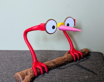 May include: A red, 3D printed bird with large white eyes and yellow glasses perched on a brown branch. The bird has a pink beak and red feet.