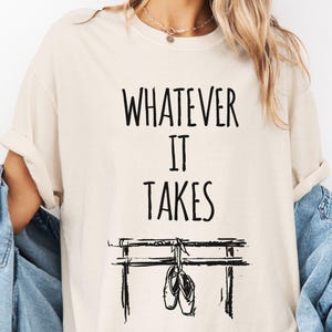 Może przedstawiać: Kremowa koszulka z czarnym napisem "WHATEVER IT TAKES". Poniżej czarna ilustracja drążka baletowego z wiszącymi baletkami. Koszulka jest w swobodnym, wygodnym stylu.