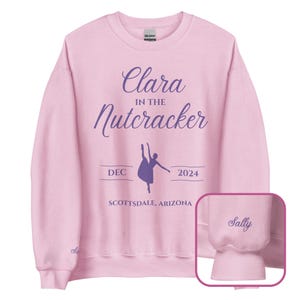 以下が含まれることがあります： 紫色の筆記体で「Clara in the Nutcracker」と書かれたピンク色のスウェットシャツ。バレリーナのシルエットと「DEC 2024 SCOTTSDALE, ARIZONA」の文字が書かれている。スウェットシャツのカフスには「Sally」という名前が紫色の刺繍で施されている。