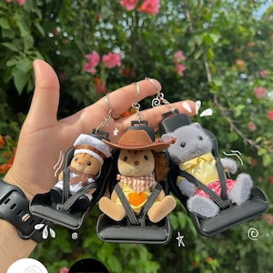 May include: Three plush toy figures, a cat, a squirrel, and a bear, are attached to keychains. Each figure is wearing a different outfit and sitting in a black plastic car seat. The keychains are attached to a silver ring.