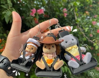 Op de afbeelding: Drie pluche figuren, een kat, een eekhoorn en een beer, zijn bevestigd aan sleutelhangers. Elke figuur draagt een andere outfit en zit in een zwarte plastic autostoel. De sleutelhangers zijn bevestigd aan een zilveren ring.