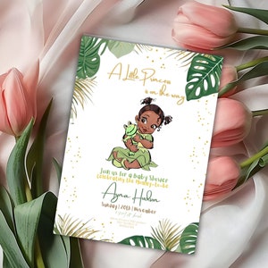 May include: A baby shower invitation with a green and gold tropical theme. The invitation features a cartoon illustration of a little girl with brown skin holding a green frog. The text on the invitation reads "A Little Princess is on the way" and "Join us for a baby shower celebrating the mommy-to-be Ayna Hudson Sunday 20th November".