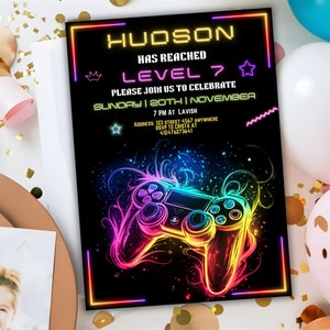 May include: A neon-colored invitation for a video game themed party. The invitation features a rainbow-colored video game controller with a black background. The text on the invitation reads "HUDSON HAS REACHED LEVEL 7 PLEASE JOIN US TO CELEBRATE SUNDAY | 20TH | NOVEMBER 7 PM AT LAVISH ADDRESS: 123 STREET 4567 ANYWHERE RSVP TO CRISTA AT 412476273641".