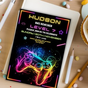 May include: Neon gamer birthday party invitation with a black background and a colorful video game controller graphic. The text reads "HUDSON HAS REACHED LEVEL 7 PLEASE JOIN US TO CELEBRATE SUNDAY | 20TH | NOVEMBER 7 PM AT LAVISH ADDRESS: 123 STREET 4567 ANYWHERE RSVP TO CRISTA AT 412476273641"