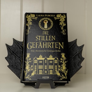 Op de afbeelding: Een zwart hardback boek getiteld "Die Stillen Gefährten" van Laura Purcell. Het boek is een Victoriaans spookverhaal en heeft een gouden illustratie van een huis met een sleutelgat op de omslag.