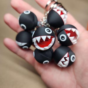 May include: A collection of black, round keychains with a cartoonish design. Each keychain features a red mouth with white teeth and white eyes. The keychains are attached to a silver chain.