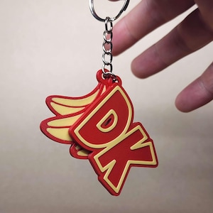 Puede incluir: Un llavero rojo y amarillo con las letras "DK" y un racimo de plátanos. El llavero está unido a un anillo y una cadena de metal plateado. El diseño es en 3D.