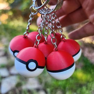 May include: A set of red, white, and black ball-shaped keyrings. Each keyring has a red top, a white bottom, and a black band with a circular black detail. The keyrings are connected to silver key rings by small chains.