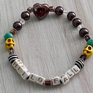 May include: A bracelet with a purple heart charm, black and white striped beads, and white beads that spell "HELLO FALL" with yellow skull beads and green accents.