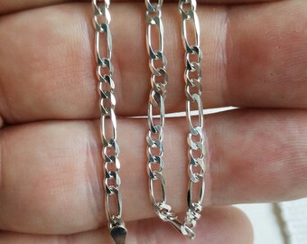 May include: A silver chain necklace with a lobster clasp closure. The chain is a figaro style with alternating flat and round links.