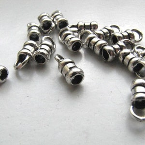 May include: A collection of silver-colored metal crimp bead tubes with a loop at one end. The tubes have a ridged design and are scattered on a white surface. These beads are used for jewelry making.