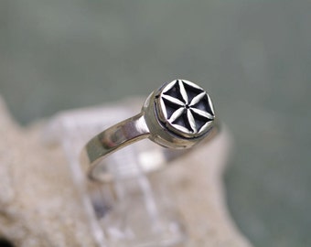 Puede incluir: Un anillo de plata con un diseño geométrico en el centro. El diseño es una flor de ocho pétalos.