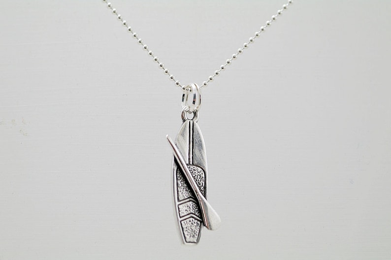 May include: A silver necklace with a pendant shaped like a paddleboard with a paddle. The pendant has a textured surface.