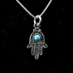 May include: A silver necklace with a hamsa hand charm featuring a blue turquoise stone.