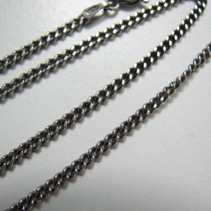 Puede incluir: Un collar de cadena de plata con un estilo de eslab&oacute;n curb. La cadena est&aacute; colocada plana sobre una superficie blanca.