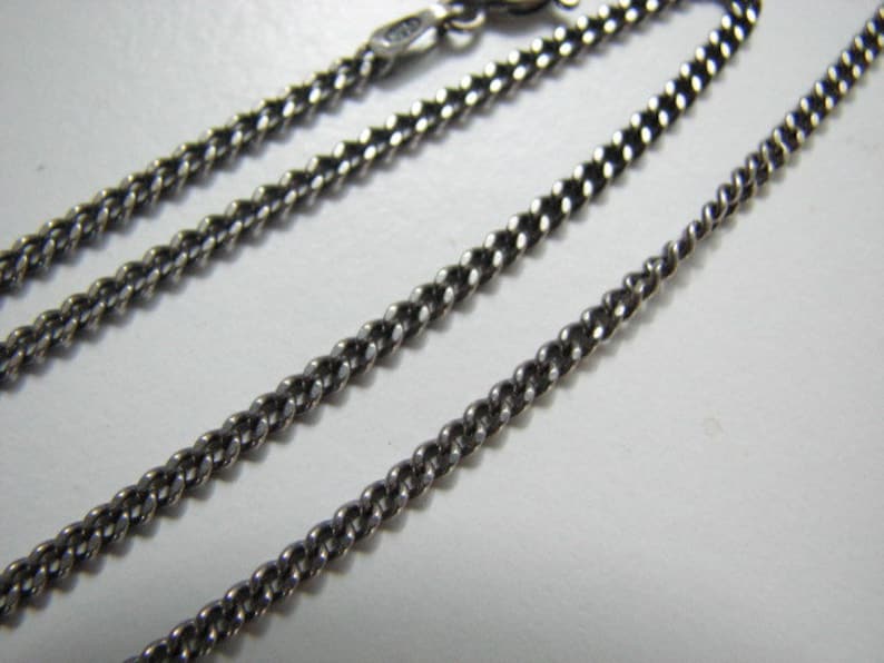 Puede incluir: Un collar de cadena de plata con un estilo de eslab&oacute;n curb. La cadena est&aacute; colocada plana sobre una superficie blanca.