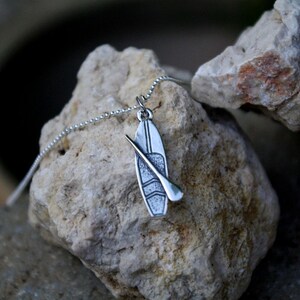 May include: A silver necklace with a pendant shaped like a paddleboard and paddle. The pendant is textured and has a detailed design.