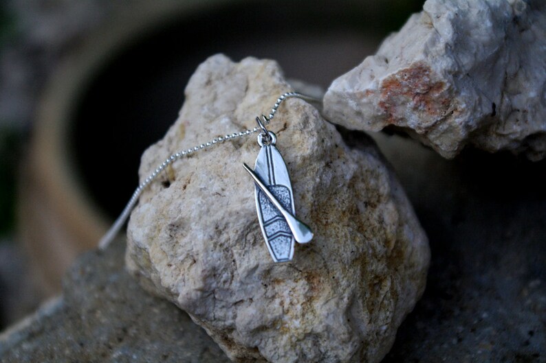 May include: A silver necklace with a pendant shaped like a paddleboard and paddle. The pendant is textured and has a detailed design.