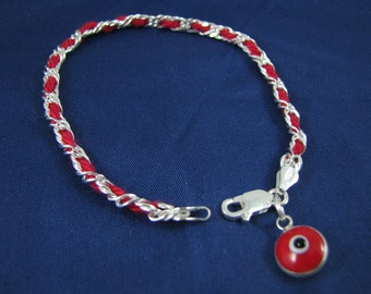 Pode incluir: Um bracelete vermelho e prateado com um amuleto prateado em forma de olho azul mau-olhado. O bracelete é feito de um cordão vermelho trançado com um fio prateado tecido através dele.