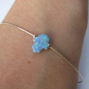 May include: A delicate silver chain bracelet with a blue opal hamsa charm. The hamsa is a symbol of protection and good luck.