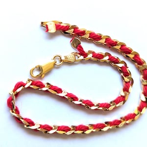 Puede incluir: Una pulsera de cadena dorada con un diseño de cordón rojo tejido. La pulsera presenta un cierre de mosquetón y se muestra sobre un fondo blanco. La joya es un accesorio elegante.