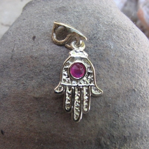 May include: A gold-coloured Hamsa hand pendant with a central red gemstone. The pendant features detailed patterns and a loop for a chain. The Hamsa hand is a symbol of protection and good fortune.