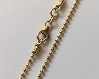 May include: A close-up shot of a gold-coloured ball chain necklace with a lobster clasp. The chain is made up of small, evenly spaced spheres. The clasp and connecting elements are also gold-toned, creating a cohesive and elegant look.