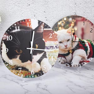 Peut inclure: Deux horloges murales rondes avec des motifs de chat noir et blanc et de chat blanc avec un pull rouge et vert. Les horloges ont des aiguilles noires et des chiffres blancs.