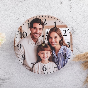 May include: A round wooden clock with a family photo printed on the face. The clock has black numbers and hands. The photo shows a family of three: a man, a woman, and a young girl.
