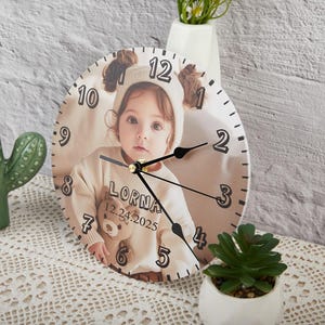 May include: A white round clock with a photo of a young girl wearing a headband and a white shirt. The clock face has black numbers and hands. The text "LORNA 12.24.2025" is printed on the clock face.