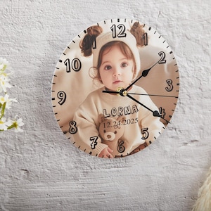 May include: A round, wooden clock with a photo of a young girl wearing a white hat and holding a stuffed animal. The clock face has black numbers and hands. The text "LORNA 12.24.2025" is printed below the photo.