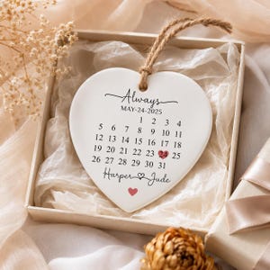 Puede incluir: Un adorno de cerámica blanco en forma de corazón con el texto Always, la fecha May-24-2025, un calendario con el día 24 resaltado por un corazón rojo, y los nombres Harper y Jude con un pequeño corazón rojo debajo, colgado de un cordel de yute.
