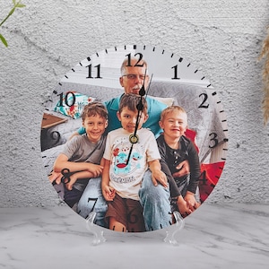 Peut inclure: Une horloge ronde avec une photo de famille, représentant un homme et trois enfants. Le cadran de l'horloge a des chiffres et des aiguilles noirs, et est présenté sur un support transparent. Le fond est un mur gris clair.