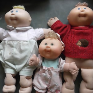 May include: Three Cabbage Patch Kids dolls, one wearing a white onesie, one wearing a pink and white dress, and one wearing a red sweater with a brown hedgehog design.