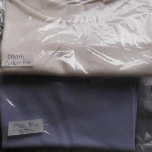 Peut inclure: Deux morceaux de tissu, l'un crème et l'autre bleu clair, sont enveloppés dans du plastique. Le tissu est étiqueté "Cream Cotton Rib" et "Med Blue Cotton".