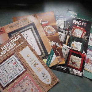 May include: Five cross stitch pattern books with colorful covers. The covers feature images of a red cardinal, a basket, a bus, a house, and a heart. The titles of the books are "Family Heritage Samplers", "Bag It", "Noah's Ark", "Impressions", and "Honey Creek Collection".