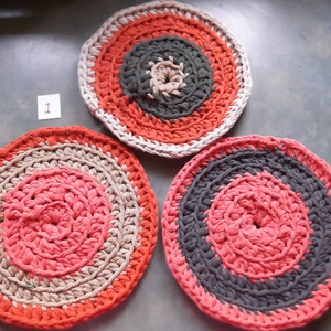 May include: Three crocheted round mats in orange, pink, gray and beige colors. The mats are made with thick yarn and have a circular pattern.
