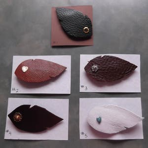 Op de afbeelding: Vijf lederen veervormen met verschillende kleuraccenten. Elke veer is genummerd met een witte sticker. De veren bevinden zich op een witte achtergrond.