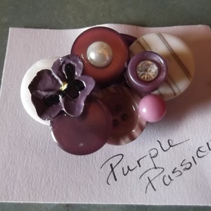 Può includere: Bottoni viola, bianchi e rosa disposti a forma di fiore con un fiore viola al centro. I bottoni sono su una carta bianca con il testo "Purple Passion" scritto in nero.
