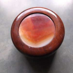 Puede incluir: Una caja de madera redonda con tapa que presenta una incrustación circular de ágata. El ágata es una mezcla de blanco, naranja y rojo.