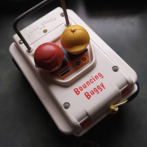 Puede incluir: Un coche de juguete de plástico blanco con una base azul. El coche tiene una figura roja y amarilla en el asiento del conductor. El coche tiene una inscripción roja que dice "Bouncing Buggy".