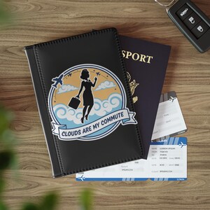 Puede incluir: Funda de pasaporte negra con un gráfico de una mujer con maleta, nubes y aviones, con el texto "Clouds Are My Commute". Se ven un pasaporte y billetes de avión. Una llave de coche está en el fondo.