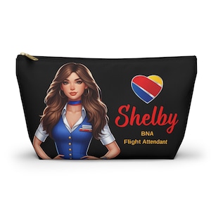 May include: Black zippered pouch featuring a stylized illustration of a flight attendant named Shelby. The design includes a heart-shaped logo with red, yellow, and blue colors, and the text "Shelby BNA Flight Attendant."