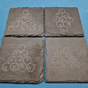 May include: Four square slate coasters with etched designs of billiard balls arranged in a triangle. Each coaster features a different arrangement of the balls, with numbers visible. The coasters are a dark gray color.