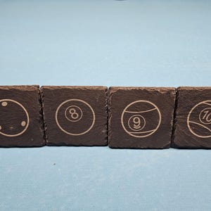 May include: Four square slate coasters with etched billiard ball designs. Each coaster features a different ball: cue ball, 8-ball, 9-ball, and 10-ball. The coasters are dark grey and sit on a light blue surface.
