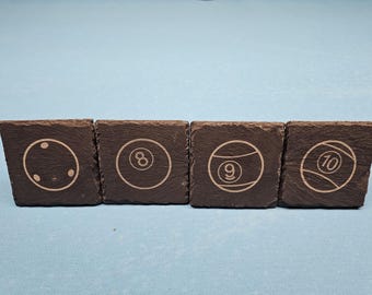 May include: Four square slate coasters with etched billiard ball designs. Each coaster features a different ball: cue ball, 8-ball, 9-ball, and 10-ball. The coasters are dark gray and sit on a light blue surface.