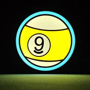 May include: A glowing, circular sign resembling a nine-ball from a pool game. The sign features a yellow and white design with a black number 9 in the center, outlined in black. The outer edge is a bright blue, set against a black background.
