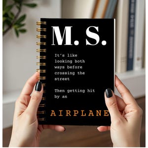 May include: Black spiral-bound notebook with gold coils. The cover displays "M.S." in white, and the text "It's like looking both ways before crossing the street. Then getting hit by an AIRPLANE" in white and orange.