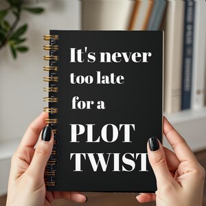 May include: A black spiral-bound notebook with gold-colored wire binding. The front cover features the white text "It's never too late for a PLOT TWIST" printed in a bold, clean font. The notebook is held upright, displaying the front cover clearly.