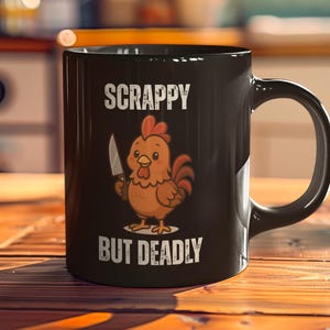 Puede incluir: Taza de cerámica negra con las palabras "SCRAPPY BUT DEADLY" y un dibujo de un pollo con un cuchillo. El pollo es marrón, naranja y rojo. La taza está sobre una mesa de madera.